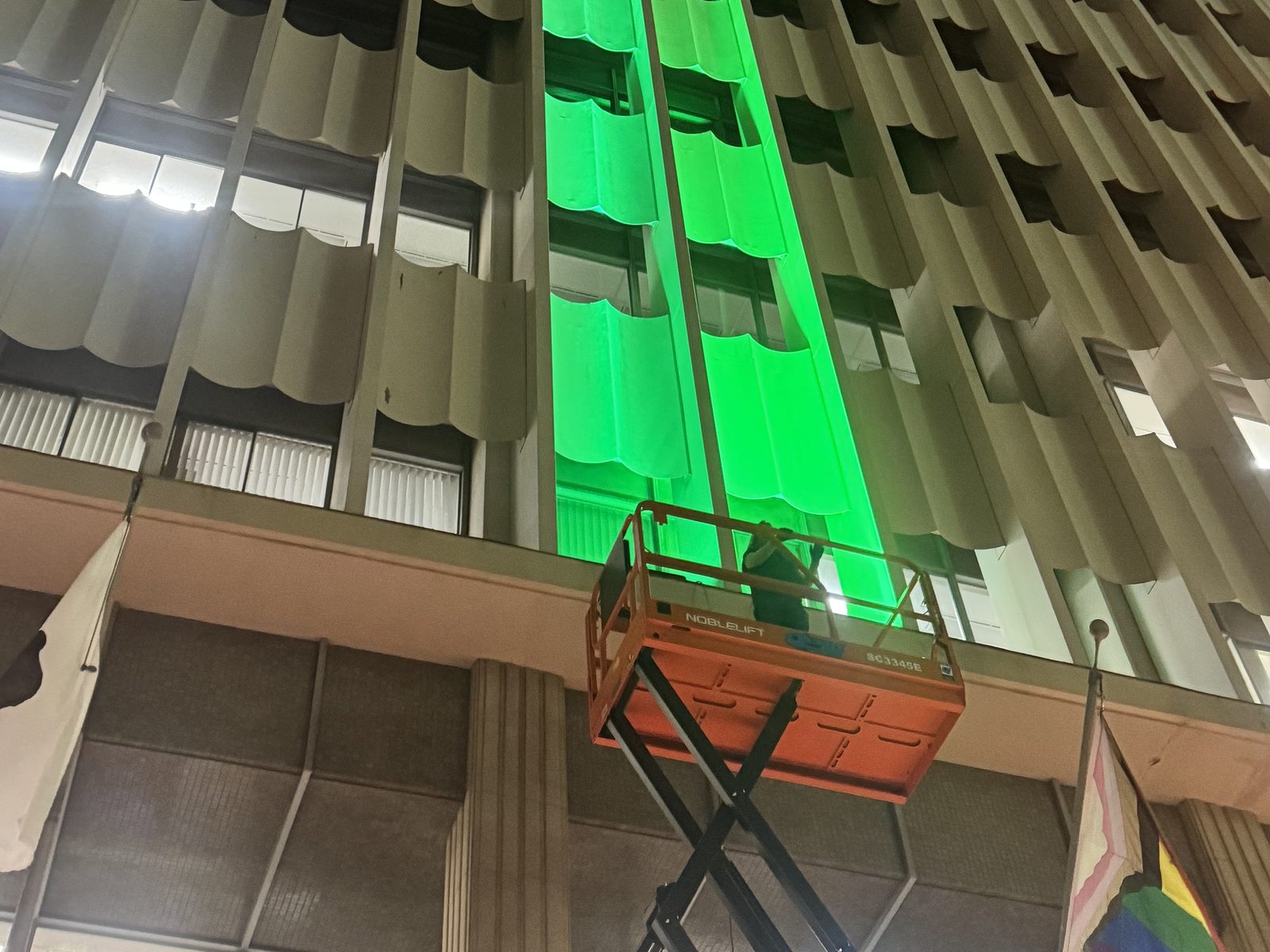 Vertical bay of the San Diego City Hall facade grazed green during an on-site lighting mockup, crew on a scissor lift below
